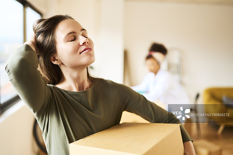一位美丽年轻女性的肖像，她长发披肩，微笑着闭着眼睛，轻触自己的头发，同时手里拿着一个搬家箱子，背景中有朋友，正面视角。图片素材