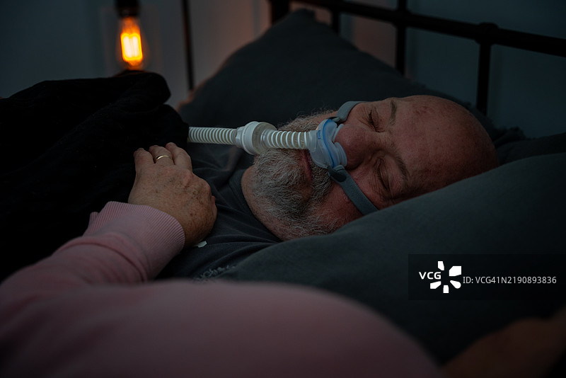 一位老年男性佩戴着CPAP（持续正压呼吸机），在被诊断为睡眠呼吸暂停后，他的妻子将手臂搁在他的胸口。图片素材