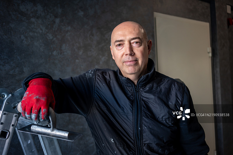 手中拿着抹子，旁边有金属楼梯，他正看向镜头，水平拍摄，家居修缮。图片素材