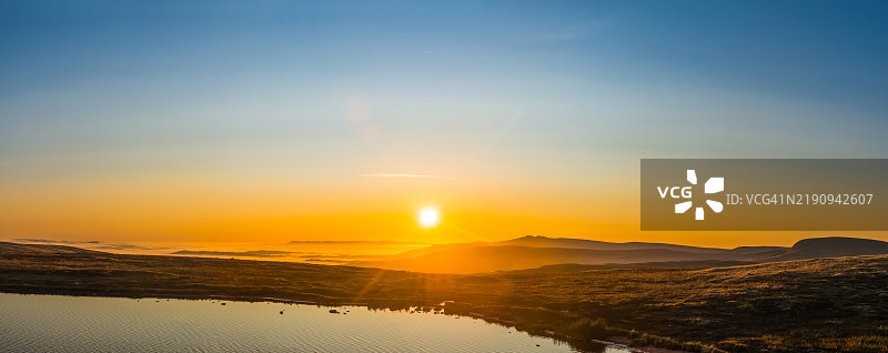 温暖的日出映照在雾气弥漫的山脊上，宁静的湖泊与荒野全景图片素材