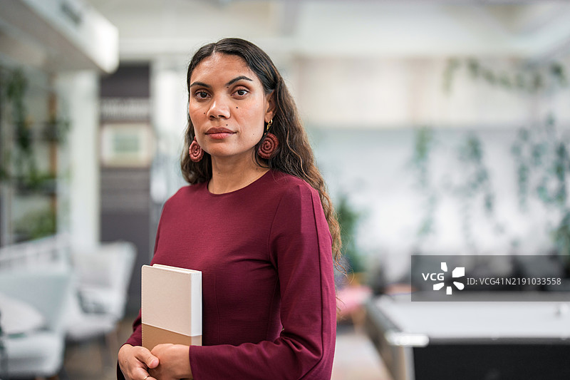 自信的年轻土著女商人在现代办公室中手持笔记本图片素材
