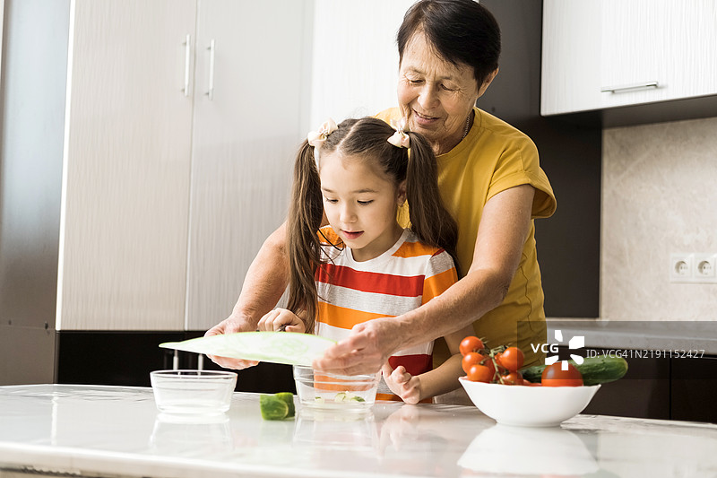 一个小白人女孩和她的祖母正在厨房里准备蔬菜沙拉。图片素材