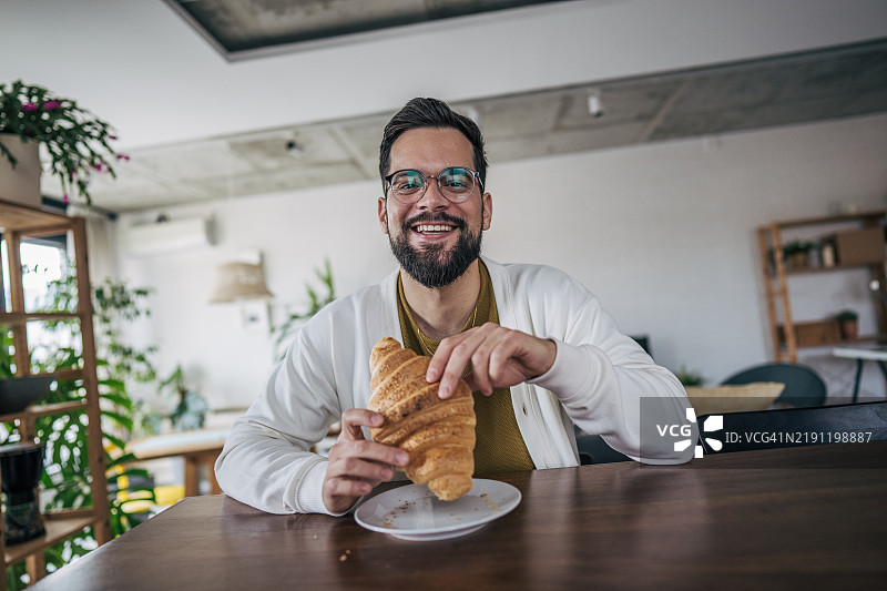 一名男子在早餐时吃着可颂面包，面带微笑地看向镜头图片素材