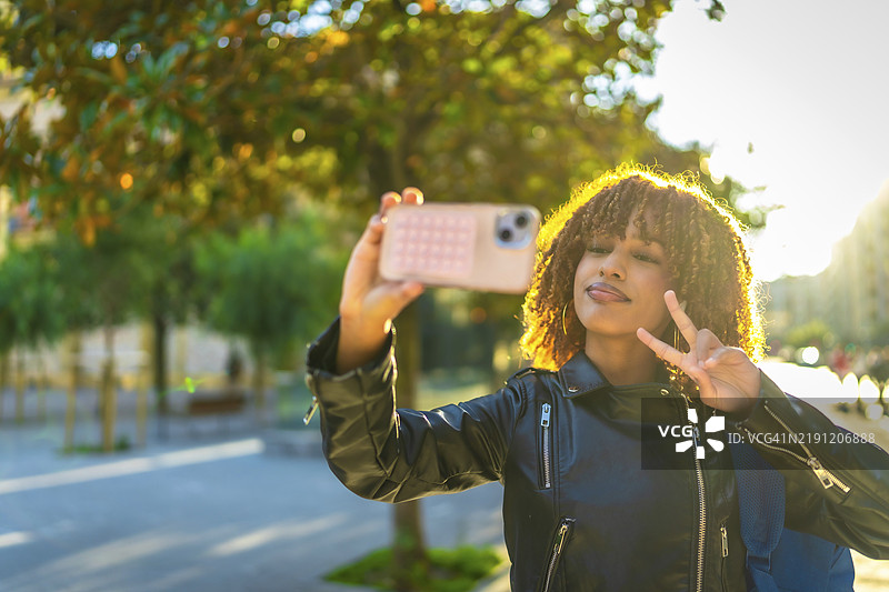快乐的拉丁裔千禧一代女性学生在日落时分于城市中用智能手机自拍并做出成功手势图片素材