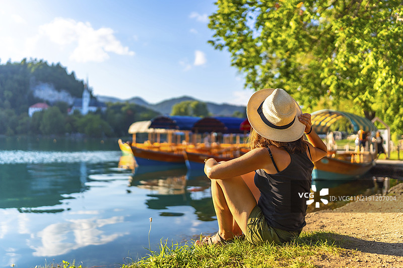 年轻女子戴着草帽坐在布莱德湖边，欣赏着传统的普莱特纳船，远处可见圣母升天教堂。图片素材