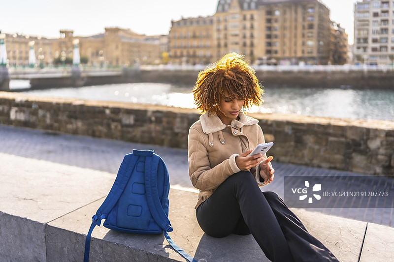 拉丁裔年轻女性坐在城市河岸长廊旁，使用手机。图片素材
