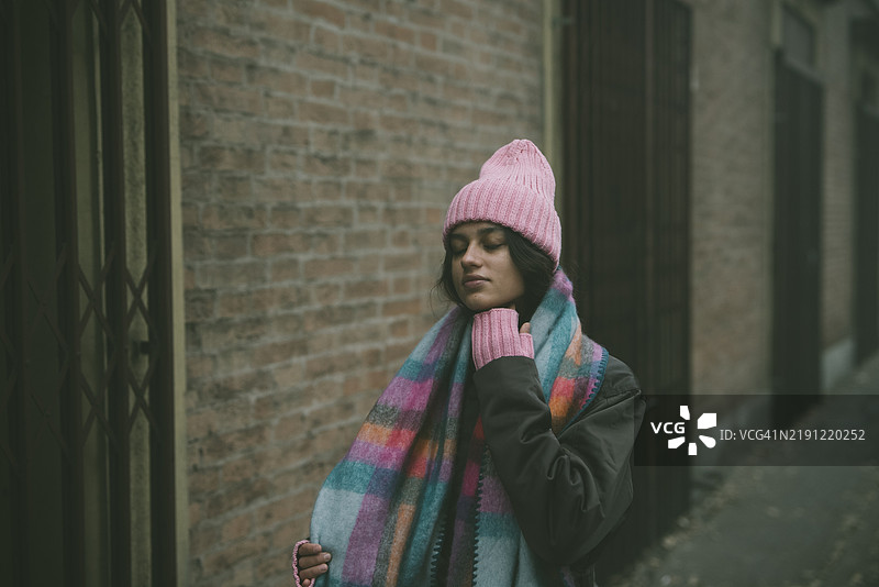 一位戴着粉色帽子和围巾的女性站在砖墙前。图片素材