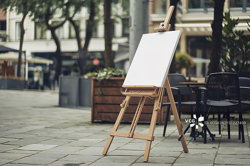 街道或咖啡区的前景中，有一个木制画架，上面放着一块空白的白色画布。图片素材