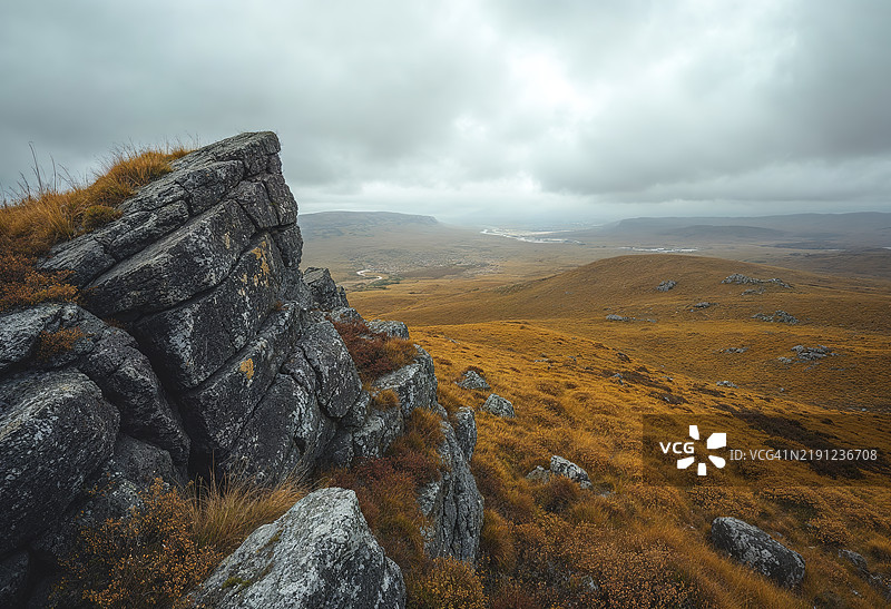 一块岩石突起俯瞰着广阔的金色荒原，天空布满戏剧性的云彩图片素材