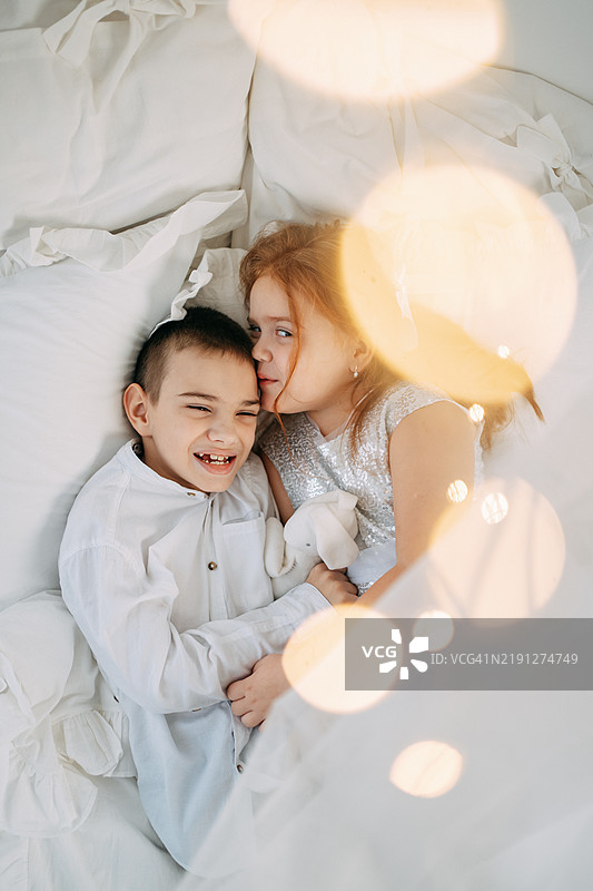 快乐的脑瘫兄弟和妹妹躺在白色床上拥抱，手里抱着一个毛绒玩具，面向镜头。图片素材