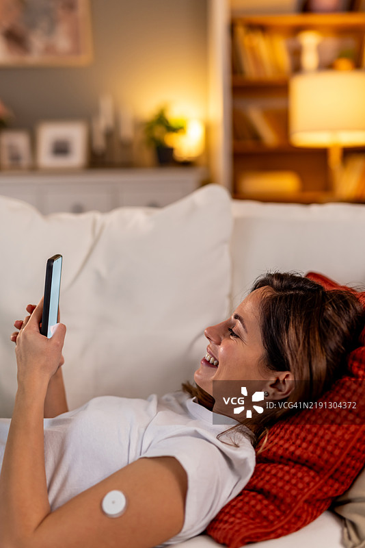 女性微笑着使用智能手机，放松地坐在沙发上，佩戴医疗设备。图片素材