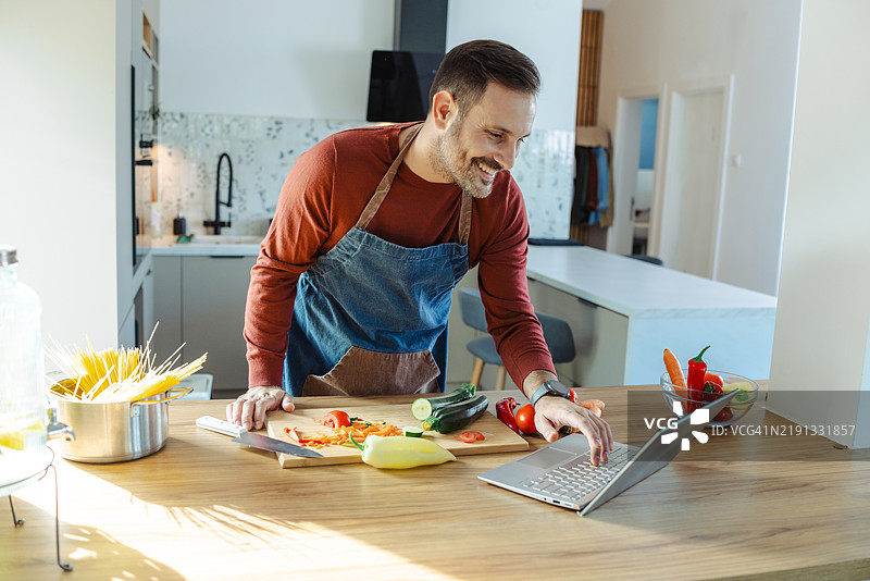男子在厨房里一边做饭一边观看笔记本电脑上的烹饪教程图片素材