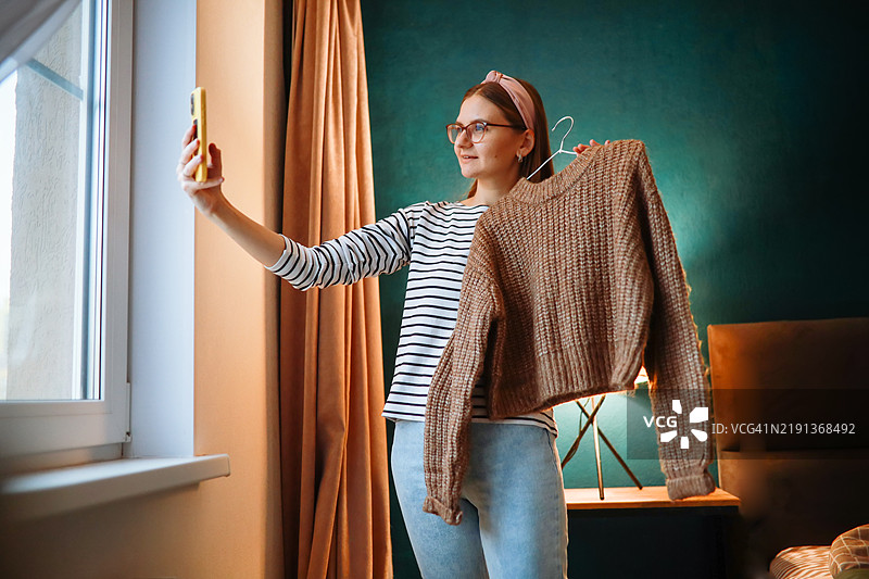 一位女性用智能手机拍摄或录制挂着的毛衣，践行有意识的消费，通过在线购物和二手衣物转售。图片素材