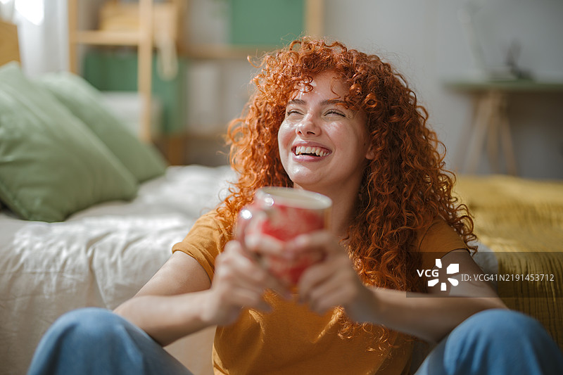 快乐的红发女性在卧室里笑着，手里拿着一个杯子图片素材