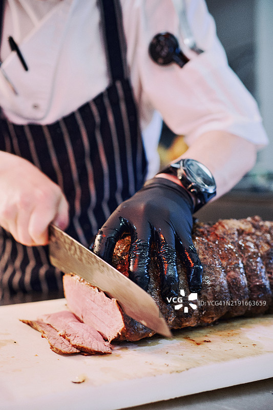 专业厨师穿着围裙，正在木砧板上切割烤好的肉块的特写垂直图像图片素材
