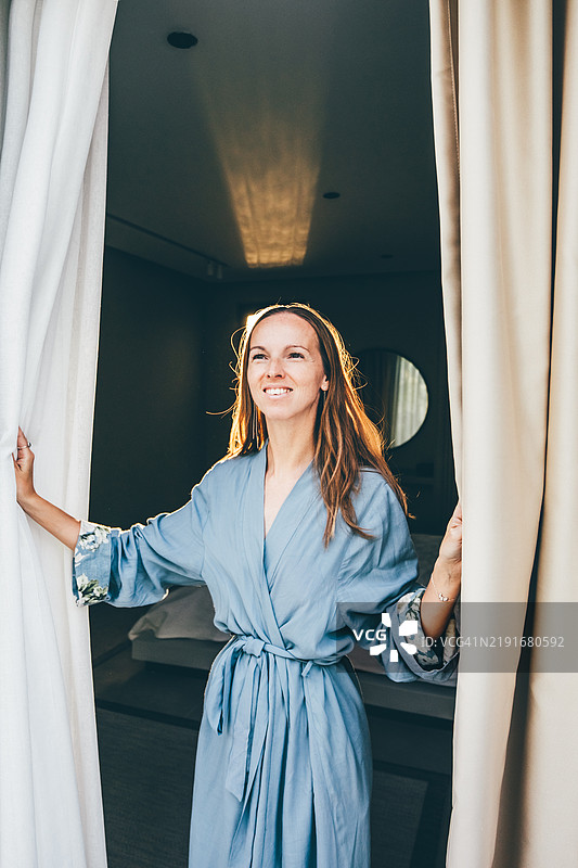 女人拉开窗帘，享受早晨大窗户外的美景。图片素材