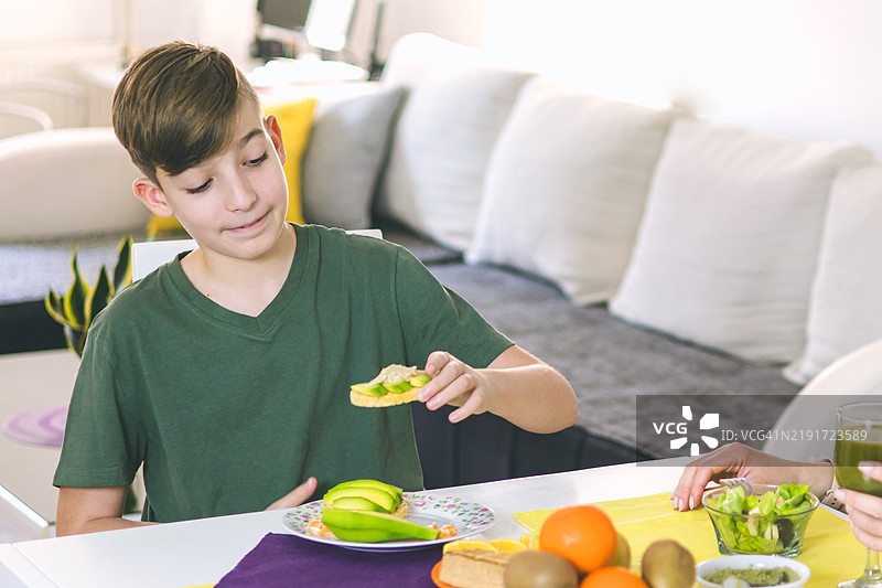母亲和儿子在家中享受准备健康早餐的时光。早餐的超级食品成分包括螺旋藻、熏豆腐、鳄梨、生菜、米饼和水果。图片素材
