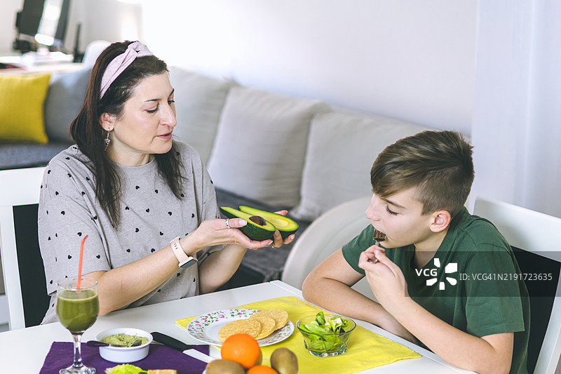 母亲和儿子在家中享受准备健康早餐的时光。早餐的超级食品成分包括螺旋藻、熏豆腐、鳄梨、生菜、米饼和水果。图片素材