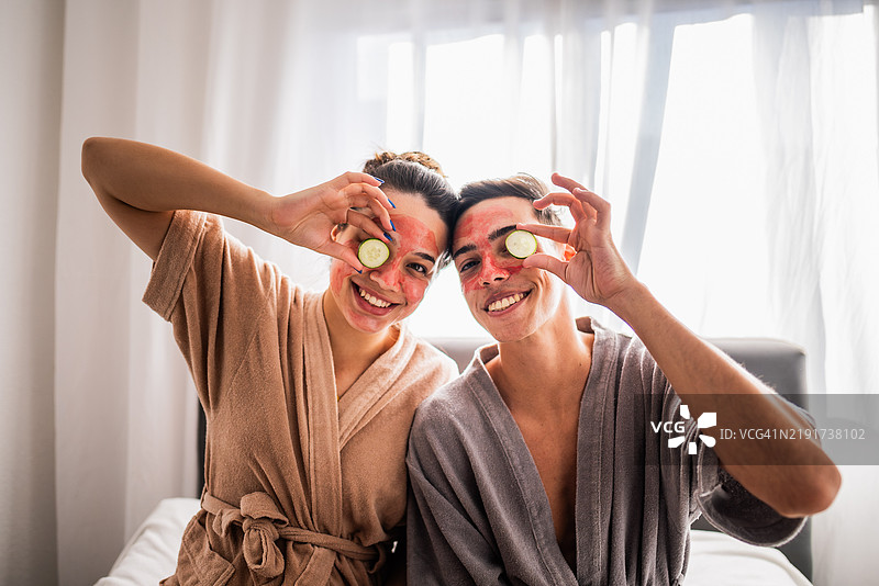 一对年轻情侣在家卧室里戴着面膜，手里拿着黄瓜片的肖像。图片素材