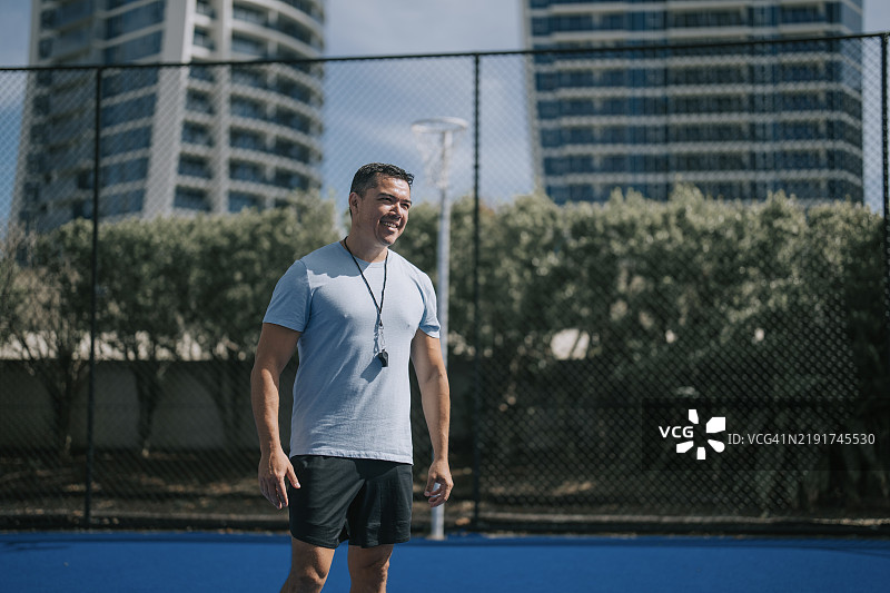 微笑着站在运动场前的澳大利亚多种族男性教练，目光投向一旁。图片素材