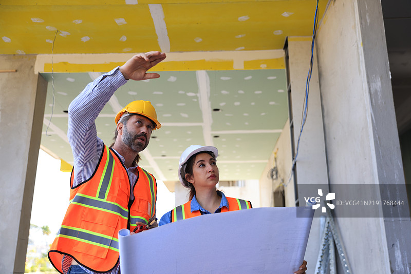 男性工程师和女性工头穿着安全背心在家居办公室施工现场一起查看蓝图图纸。图片素材