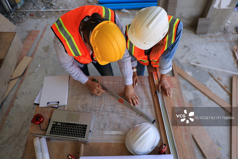 男性工程师和女性工头穿着安全背心在家居办公室施工现场一起查看蓝图图纸。图片素材