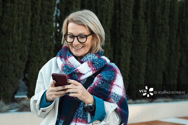 冬日里，一位成熟的金发快乐女性戴着眼镜，正在使用手机。图片素材