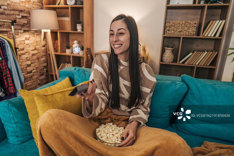 一位年轻女性坐在舒适的客厅沙发上，边吃爆米花边观看她最喜欢的电视节目。图片素材