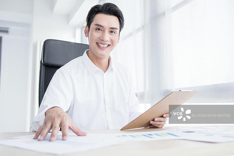 一位年轻的职业男性穿着白衬衫，坐在办公室的桌子前，正在审阅一些文件并对着镜头微笑。图片素材