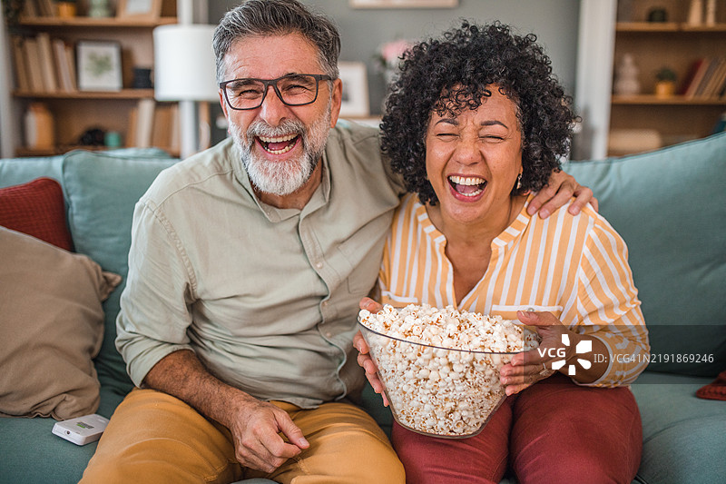 一对快乐的多种族情侣在客厅里疯狂地看电视，吃着爆米花。图片素材