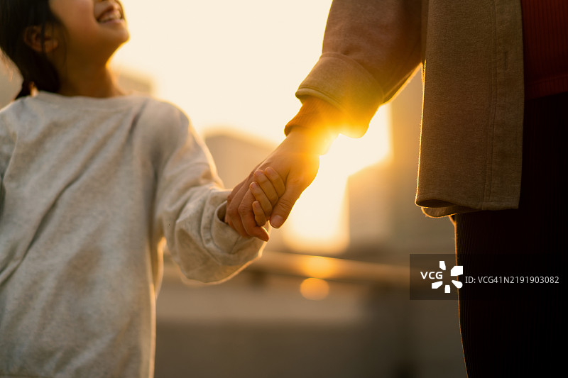 年轻的亚洲母亲与女儿手牵手走在城市中，享受着美丽金色日落下的家庭亲密时光。健康的家庭生活方式。家庭的爱、关心与团结。图片素材