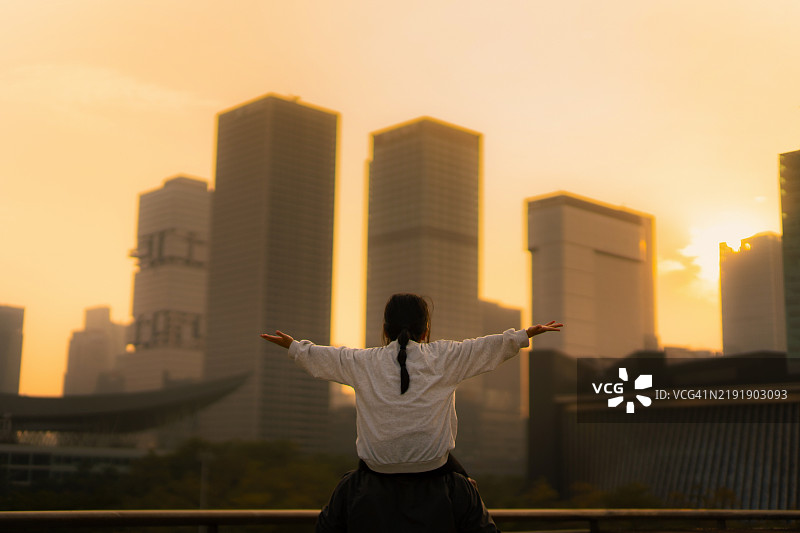 年轻的亚洲父亲将女儿抱在肩上，双臂伸展，俯瞰现代摩天大楼下的城市全景，金色日落映衬着他们。享受父女之间的亲密时光。家庭的爱与团结生活方式。图片素材