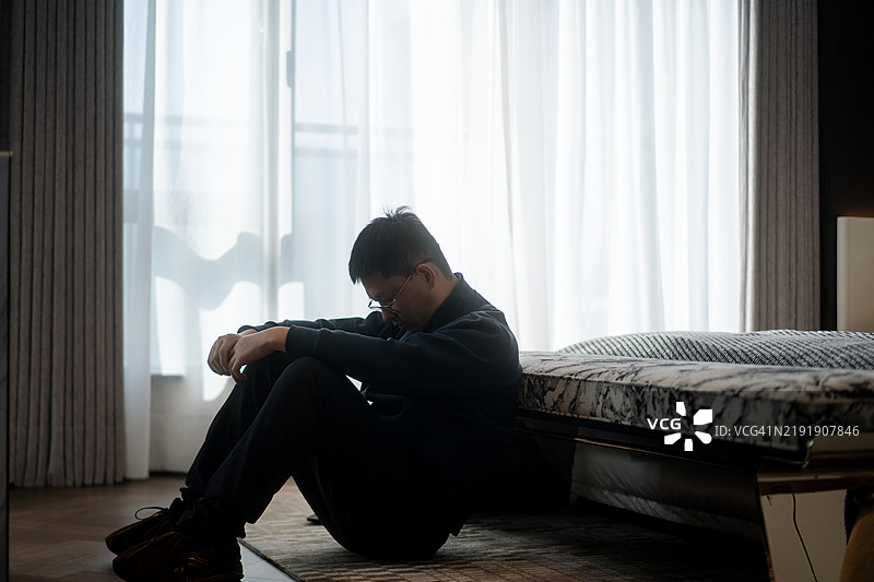 这个男人坐在床边的地上，神情沮丧。图片素材