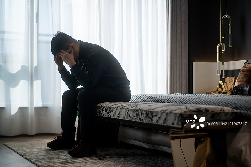 一位亚洲男子坐在床边，头痛不已。图片素材