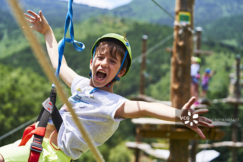 一个男孩兴奋地悬挂在滑索上，双臂伸展，背景中可以看到模糊的平台和更多的人。图片素材