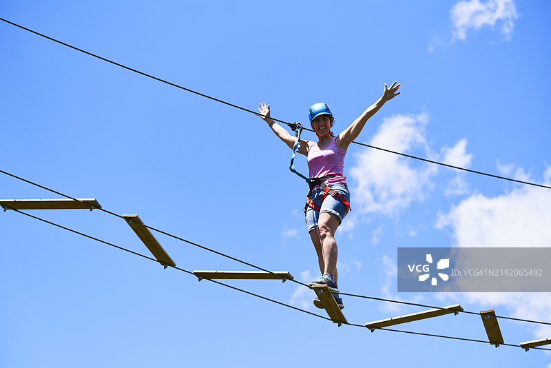 一位女性站在悬索桥上，微笑着张开双臂，面向镜头，身穿安全装备。图片素材
