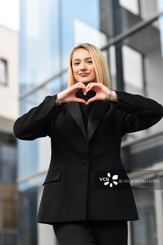 美丽的女性站在办公大楼前，用手比心，面向镜头。图片素材