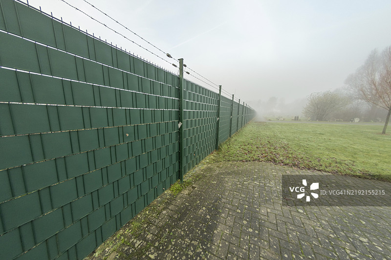 绿色塑料围栏顶部带有刺铁丝，分隔私人财产与公共空间，雾天景象图片素材