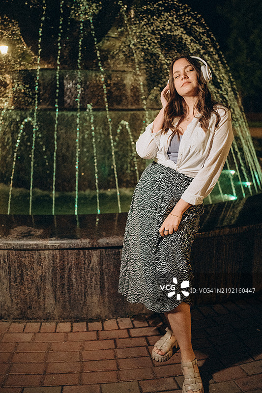 年轻女性在夜晚的喷泉旁戴着耳机听音乐图片素材