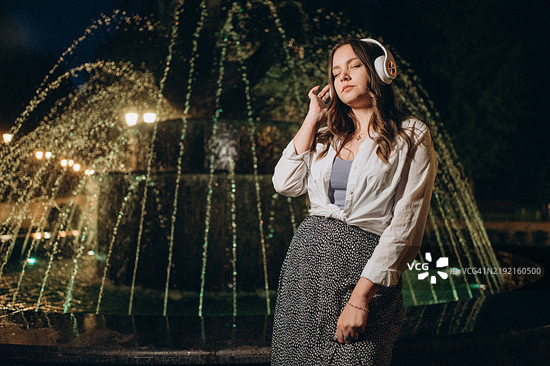 年轻女性在夜晚的灯光喷泉旁戴着耳机听音乐图片素材