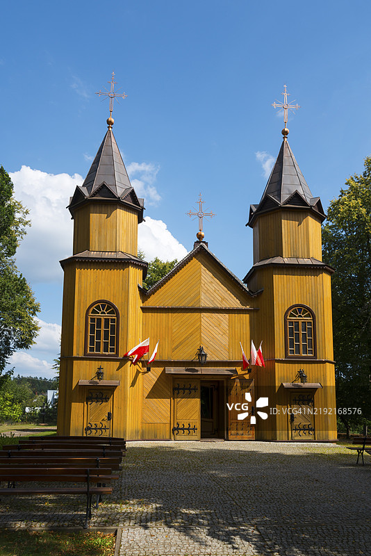 正面视图的木制教堂，带有塔楼，悬挂波兰国旗，周围环绕着树木和蓝天，圣玛丽·玛格达琳教堂，米卡绍夫卡，奥古斯托夫县，波德拉斯基省，波兰，欧洲图片素材