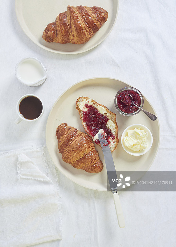 白色桌布上的可颂面包，配有黄油和果酱，旁边是咖啡和牛奶，从正上方拍摄。图片素材