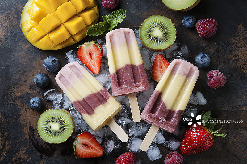 一个乡村风格的黑色背景，上面摆放着由芒果、奇异果和草莓制成的冰棒图片素材