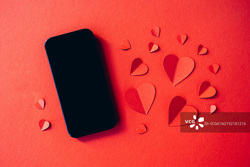 智能手机与纸质心形图案在鲜艳的红色背景上，完美适合情人节或爱情主题的设计图片素材