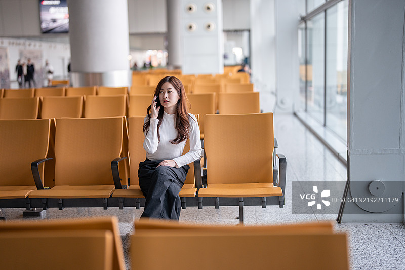 在机场候机厅等待航班的年轻亚洲女性正在使用智能手机，手里提着行李。她是一位因工作出行的亚洲女商人。科技与生活方式。旅行和度假的理念。图片素材