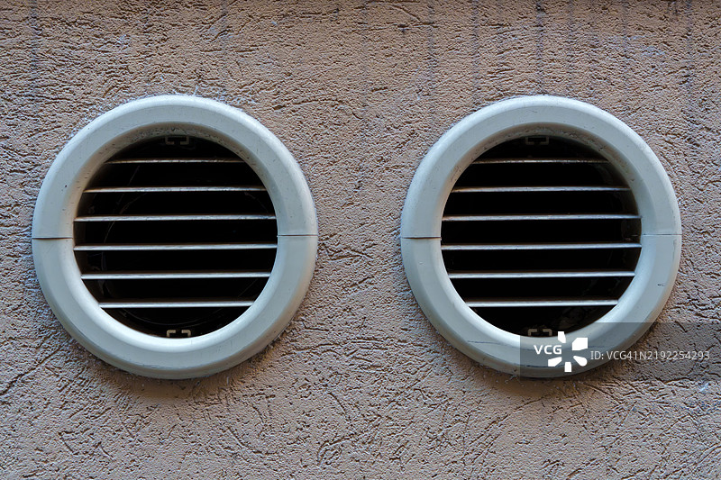 两个圆形通风格栅嵌入在一面有纹理的浅棕色墙壁中，以确保良好的空气流通和通风。图片素材