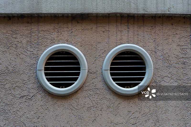 两个圆形通风格栅嵌入在一面有纹理的浅棕色墙壁中，以确保良好的空气流通和通风。图片素材