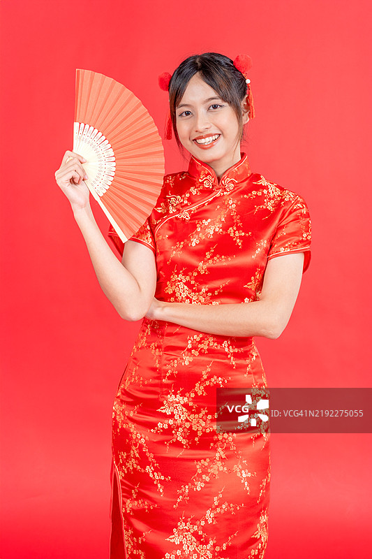 新年快乐。亚洲年轻女性穿着传统的旗袍，手持扇子，背景为红色。图片素材