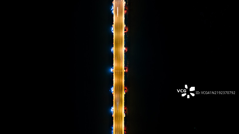 夜间对称照明下的平静水面上照亮的桥梁鸟瞰图。图片素材