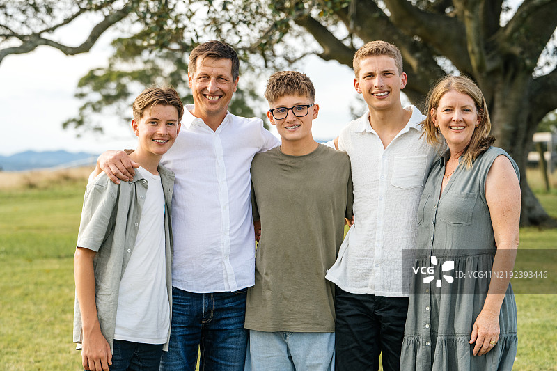 一个快乐的白人五口之家在阳光明媚的公园里微笑着，彼此靠得很近，背景是高大的树木。图片素材